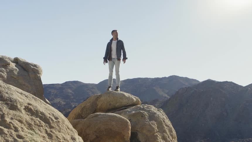 A man stands on a boulder at the very top of the mountain
