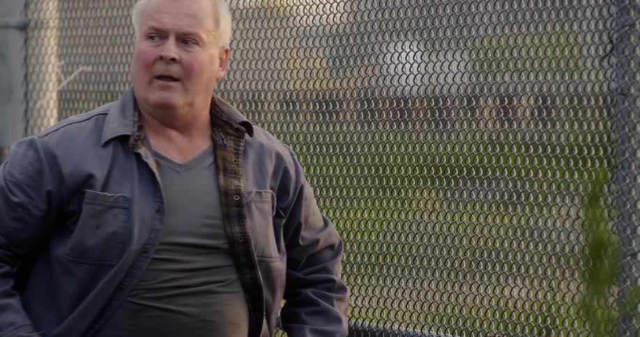 An elderly man leans against a chain link fence