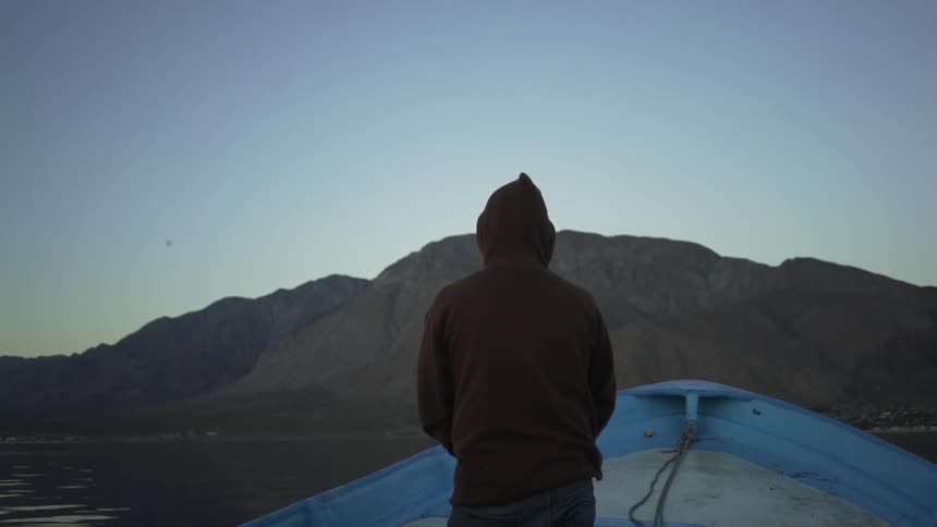 The backside of a man standing at the front of a boat looking at a mountain