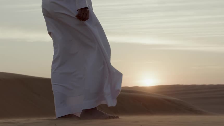 Bottom half of man in robes standing in desert at sunset