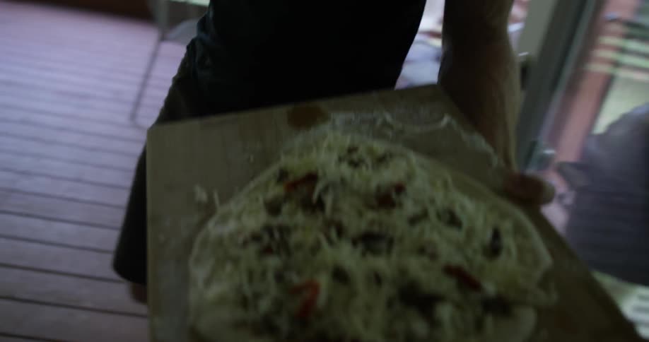 A man carries a handmade pizza to a small grill on a patio