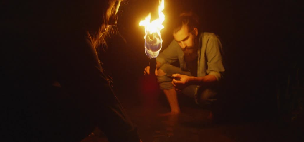 Couple Walking Through Woods Carrying A Torch Searching A Creek