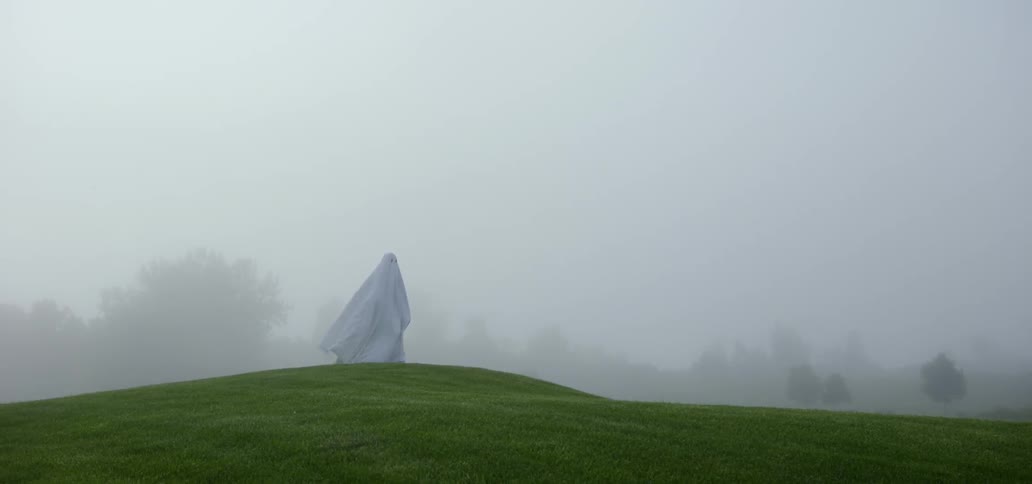 A cloaked figure walks on a grassy hill surrounded by mist