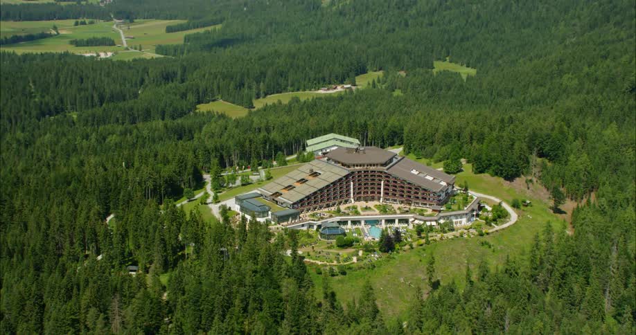 A large hotel complex sits atop a hill surrounded by forests and mountains