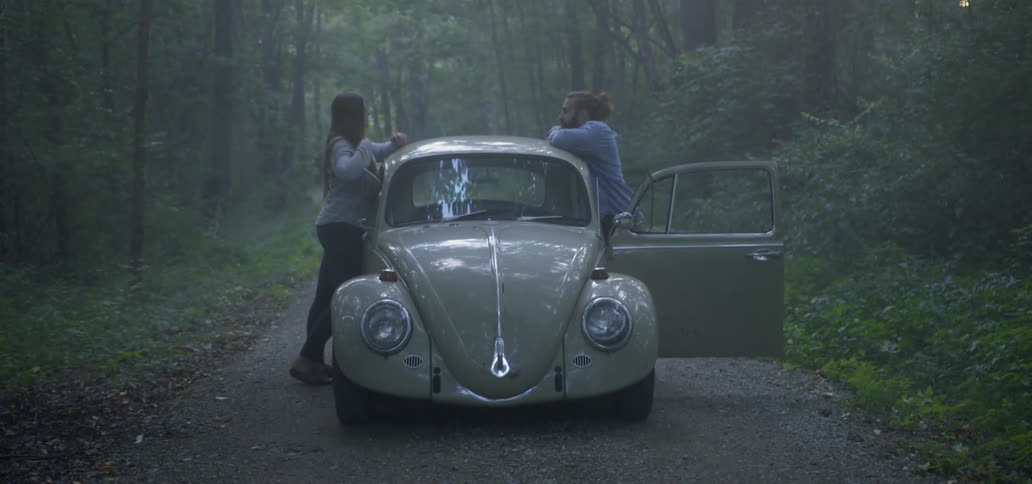 Man and woman lean against a volkswagen as it's stopped in the forest