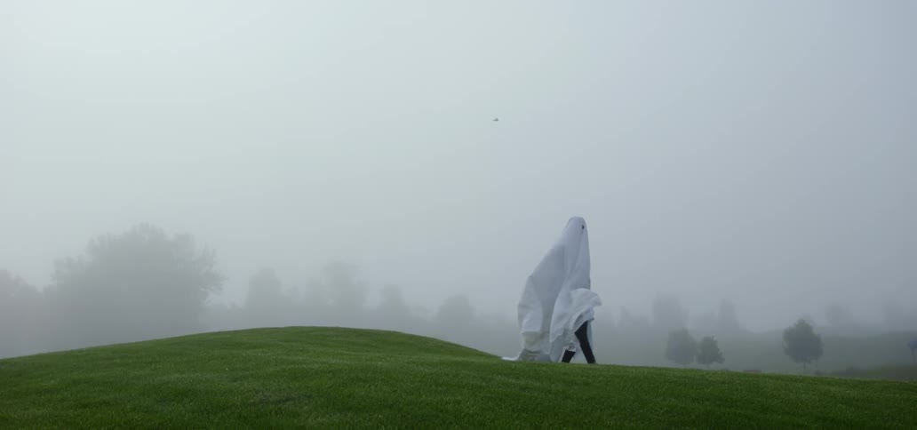 A person walks on a green hill wearing a sheet over their head