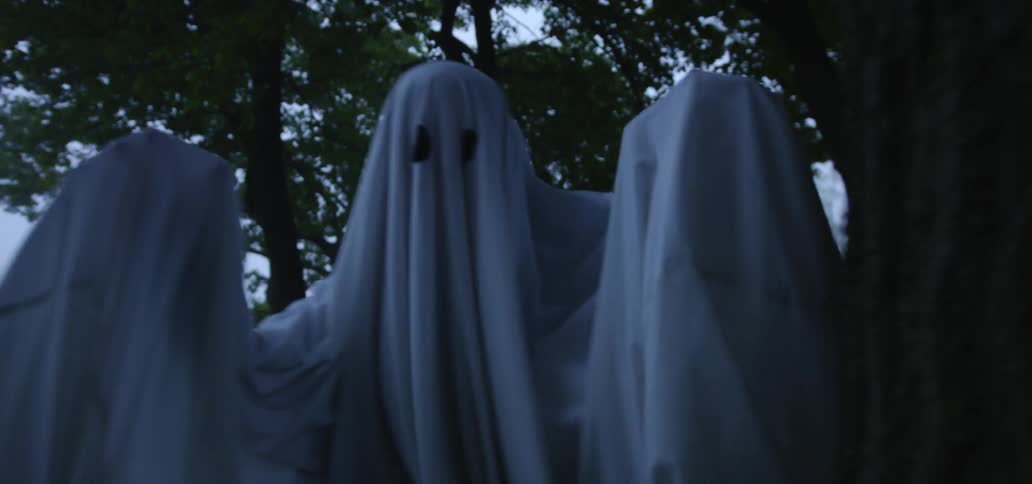 A person dressed as a ghost jumps from behind a tree at twilight