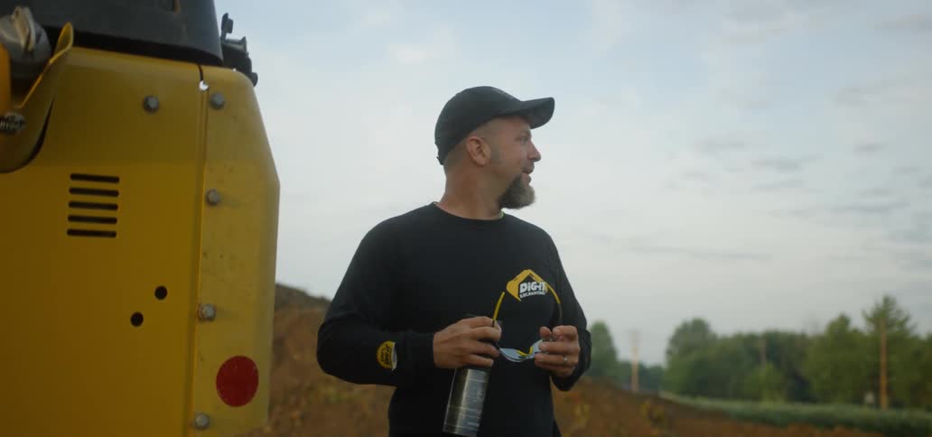 A man holds his sunglasses and a thermos while standing in front of a farm vehicle
