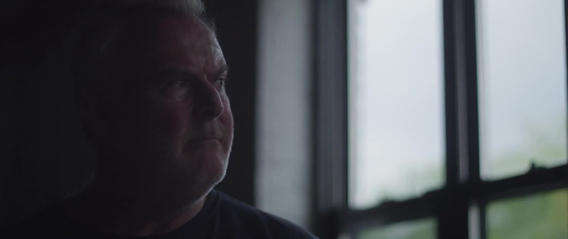 A man sitting in a window-lit room looks up and towards the windows