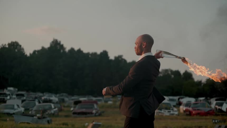 A man in a suit throws a molotov cocktail in a junkyard