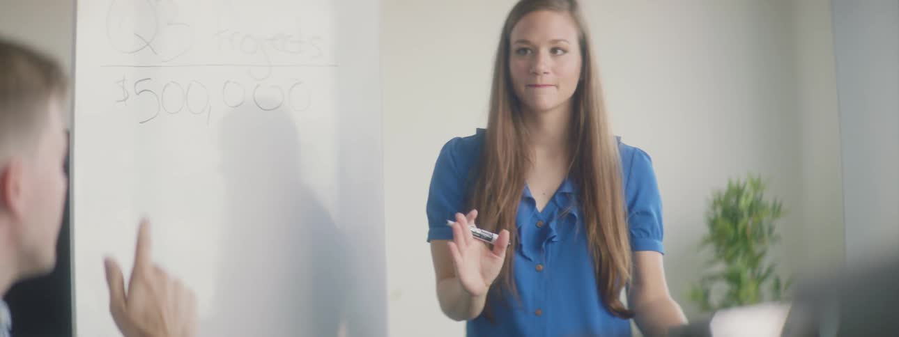 Someone types on a laptop at a meeting while a woman writes on a whiteboard
