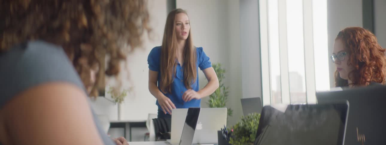 People sit in front of laptops in a boardroom as one woman stands at the head of the table