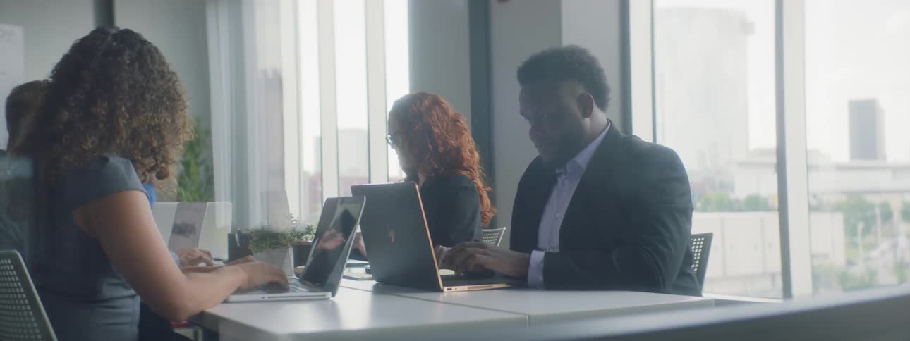 Men and women sit around a table typing on laptops