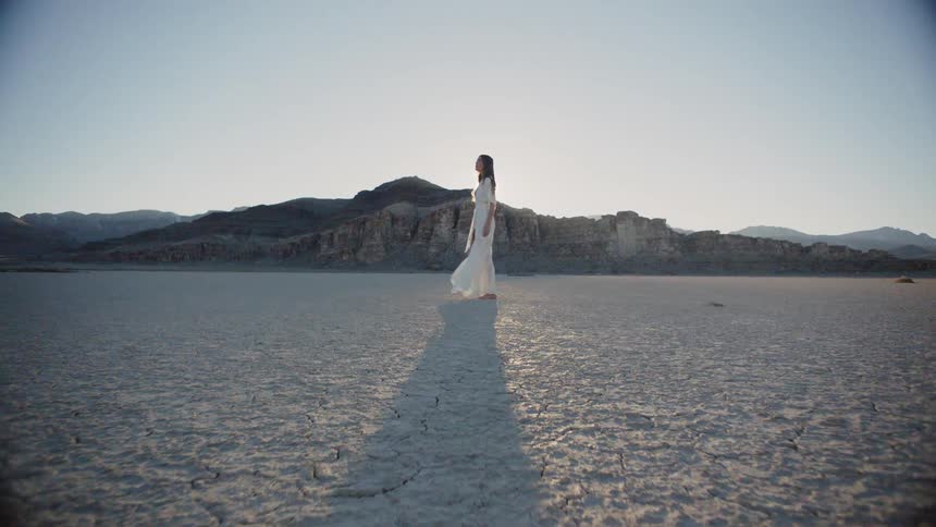 A woman in a white dress walks in a desert on a sunny day