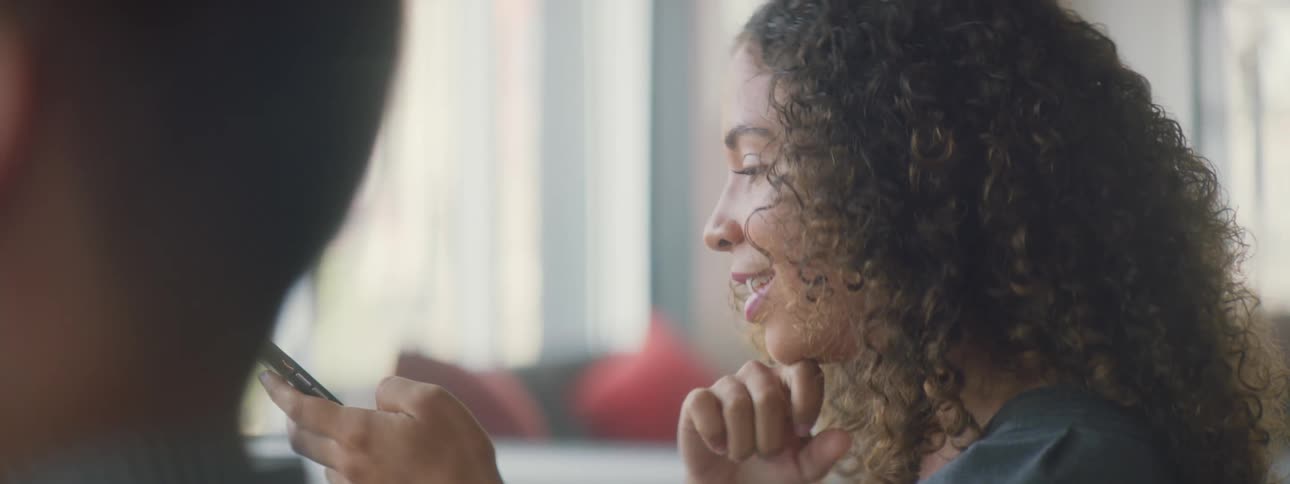 A woman smiles as she uses her phone then looks over to the person beside her