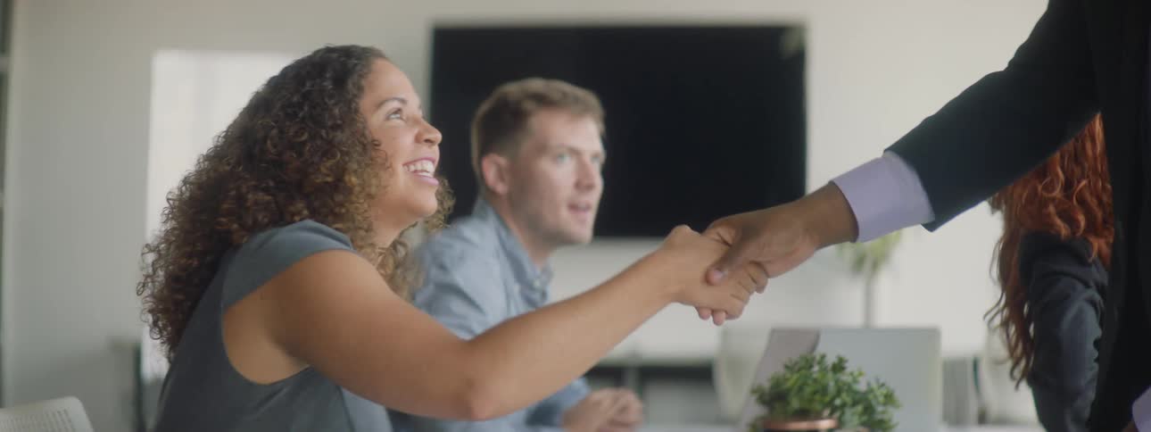 A business man reaches across a conference table to shake hands as everyone stands