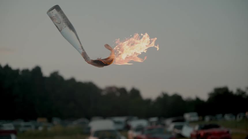 A bottle with flames coming out of it flies in the air in a grassy parking lot at sunset