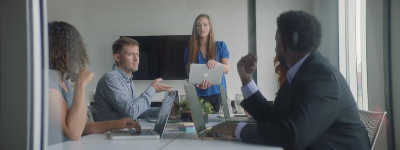 A woman stands at the head of a table full of people in a boardroom