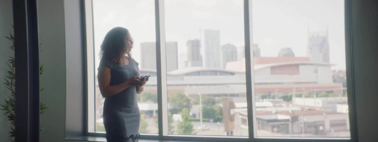 A woman types on her phone while standing in front of a large window