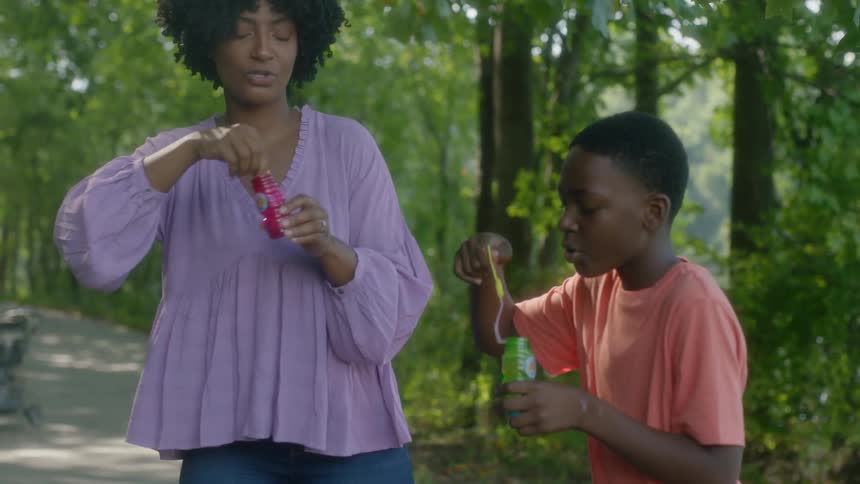 A mother and son blow bubbles while in a park with other people sitting on a bench watching