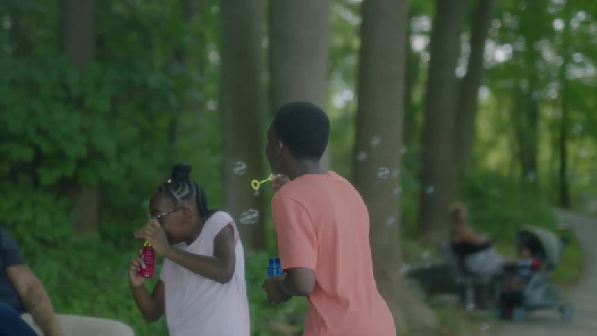 Two pre teens blowing bubbles in a park near trees and people