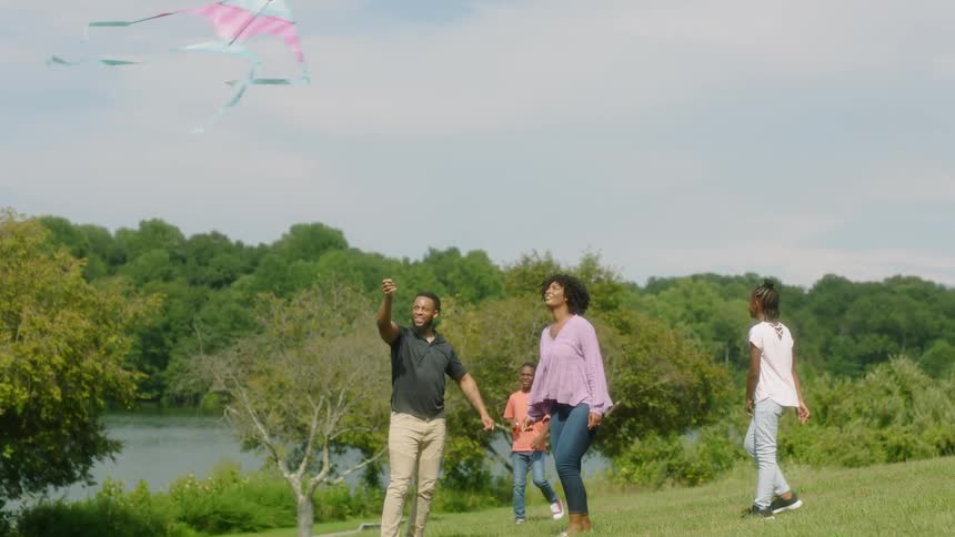 A family of four flying a kite in a park near a river and trees