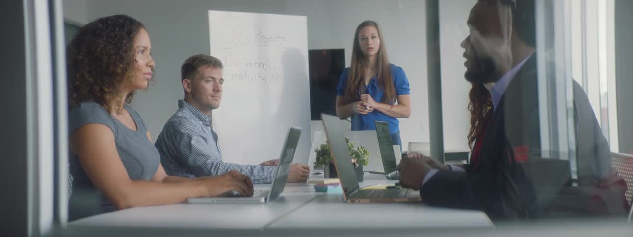A woman at a meeting writes ideas on a whiteboard as other people discuss