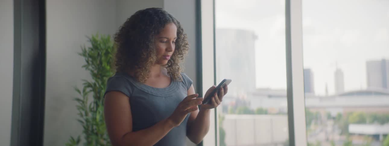 A woman scrolls on a phone while standing next to a window