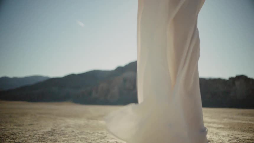 Woman wearing white dress walk barefoot in desert while the sun goes down