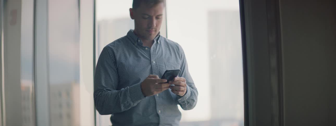 Man types on his phone in front of a large window and looks up to smile