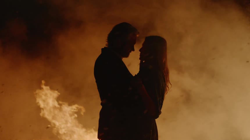 A man and woman embrace outside while smoke and ash from a fire blows past