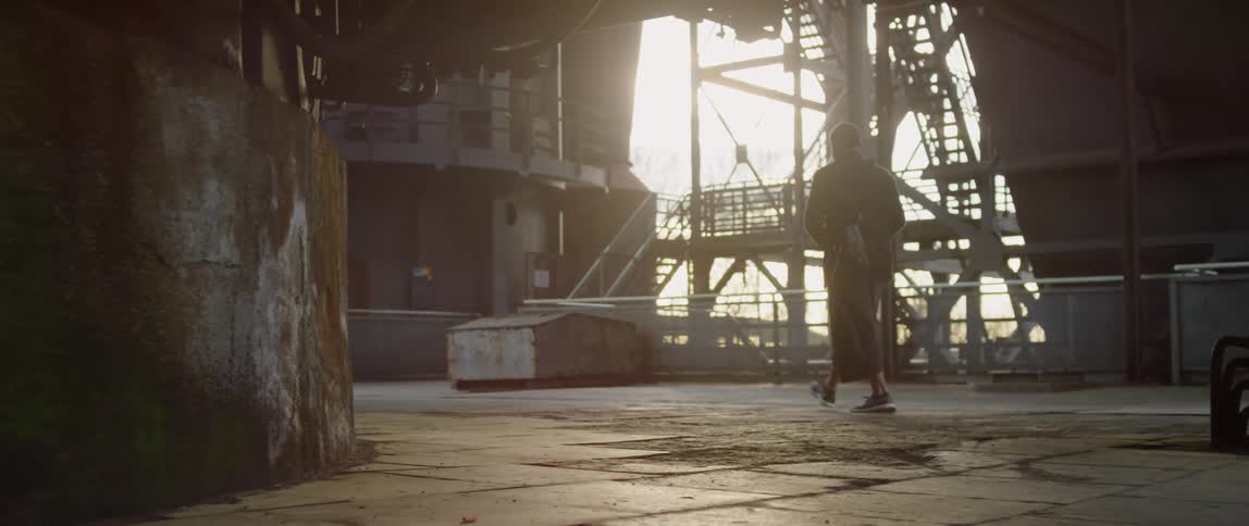 A person in loose three quarter lengths walks in an empty sunlight industrial landscape