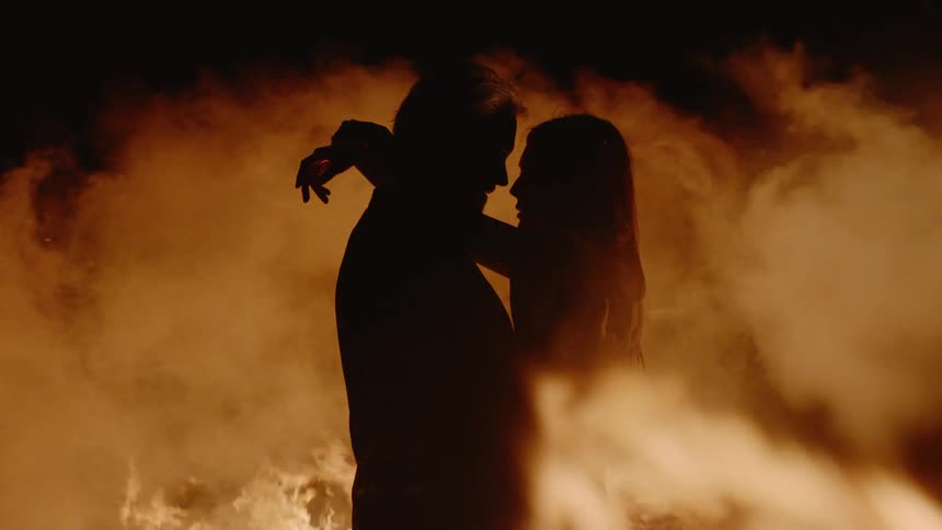 A man and woman holding each other with fire burning around them
