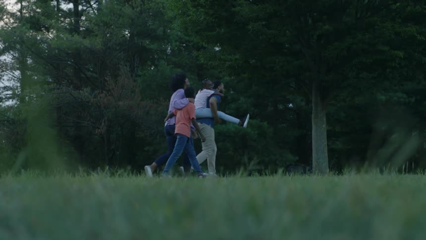 Man carries girl on shoulders while boy and woman walk behind them in forest at dusk