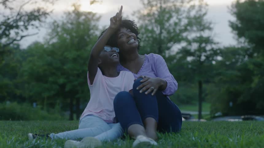 A child is sidled to a woman as they sit on the grass at sunset and laugh