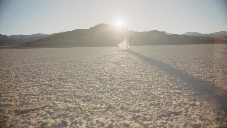 A woman in a white dress walks in a desert on a sunny day