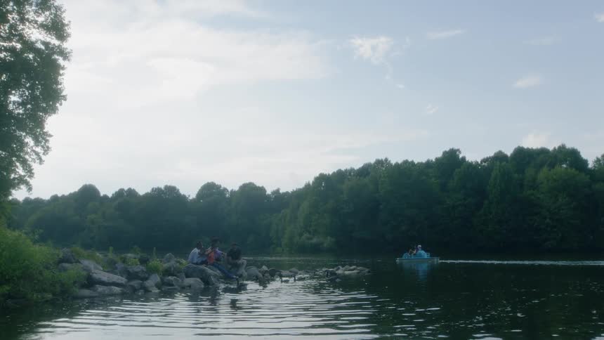 A group of people sit on rocks by a lake as a boat slowly passes by