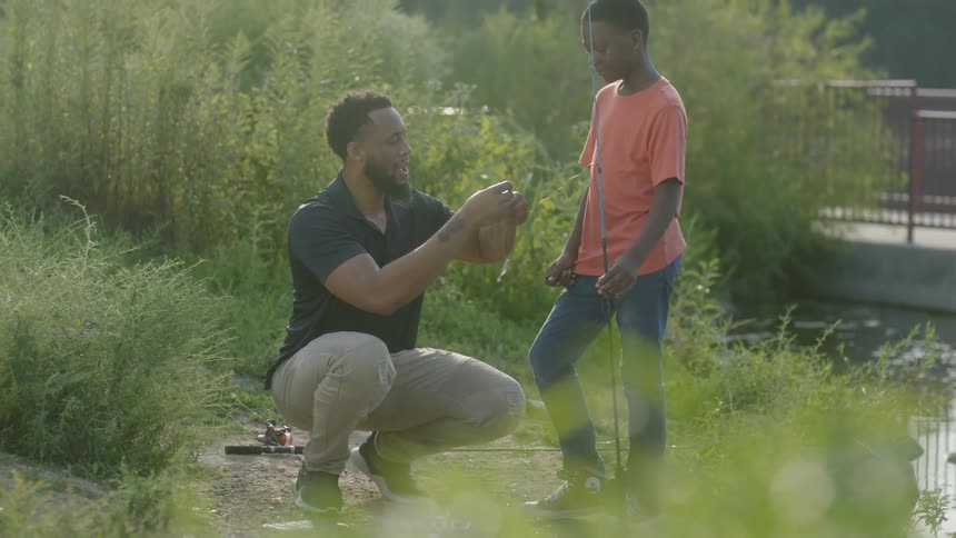 A man showing a younger man how to bait a hook near a lake