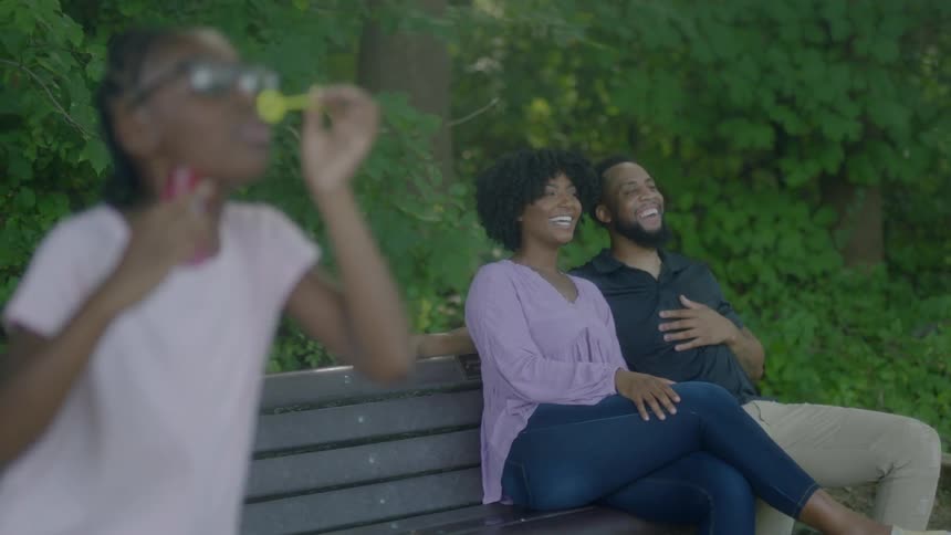 A girl blowing bubbles while her mom and dad sit laughing on a park bench
