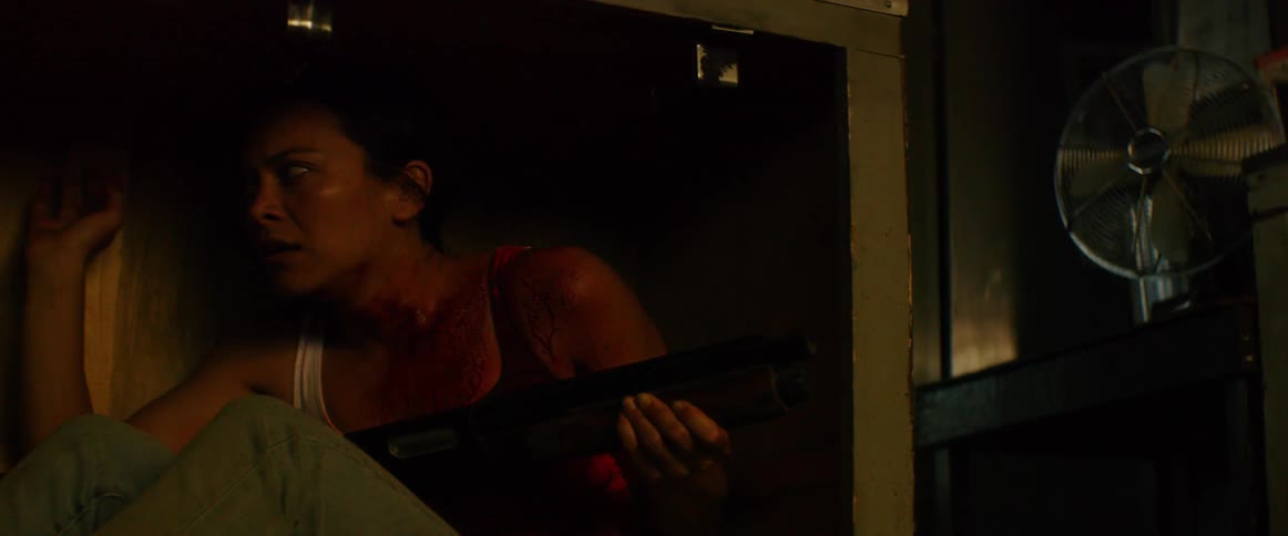 Woman covered in blood hides under furniture holding a shotgun while lights flicker