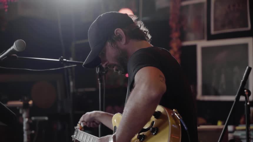 A white male dressed in black sits on a stool tuning an electric guitar with a microphone in front of him