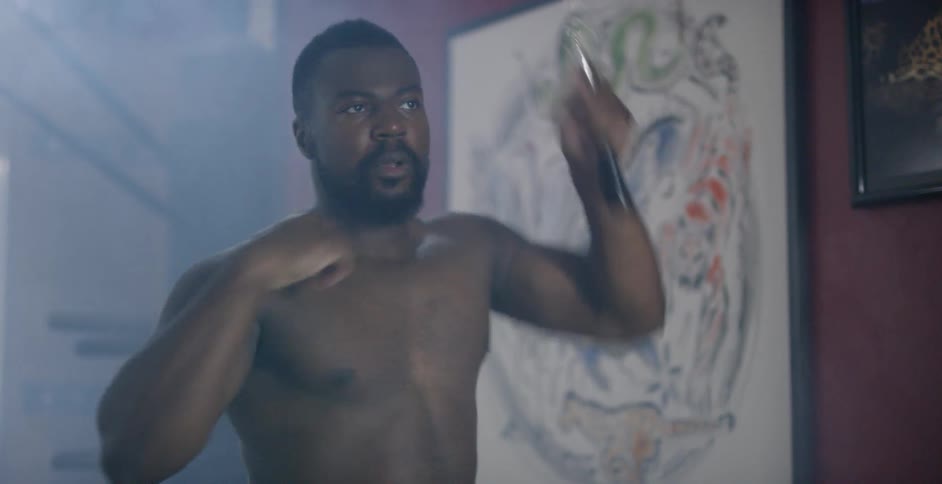 A shirtless muscled man using martial arts weapons inside of a studio