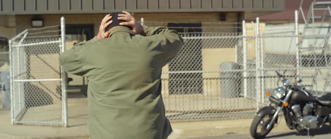 A large man outside puts on a hat and walks towards a gate and fence