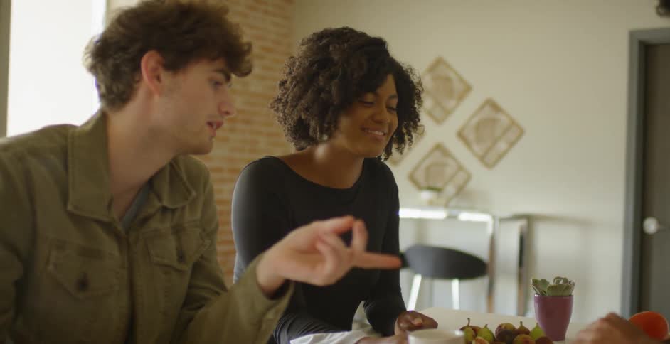 Friends sit at a  kitchen island talking and drinking as another cuts up an eggplant