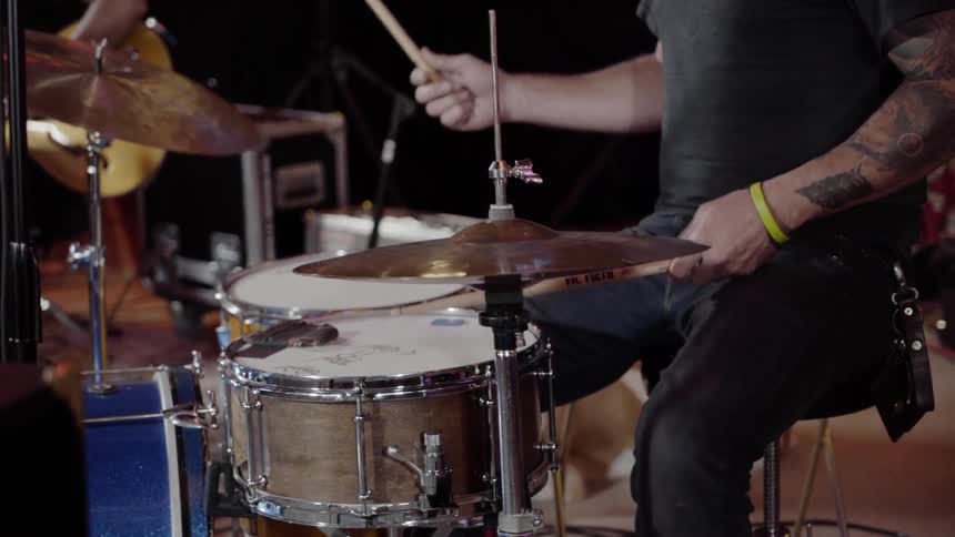 A man plays the drums as another tunes up his guitar