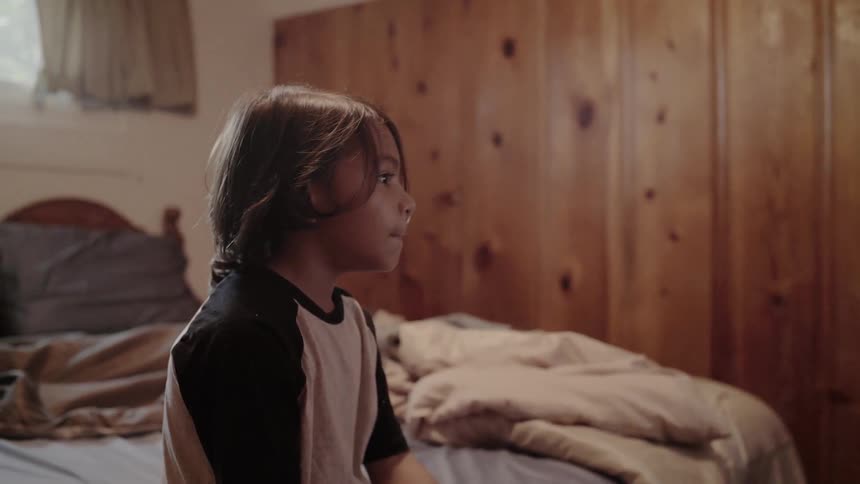 Boy sits on the side of a bed while a tv flickers in front of him