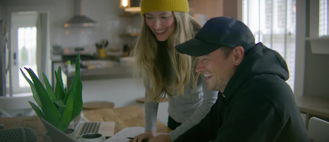 A man sitting in a chair and a woman standing in the kitchen laugh as they look at a laptop screen
