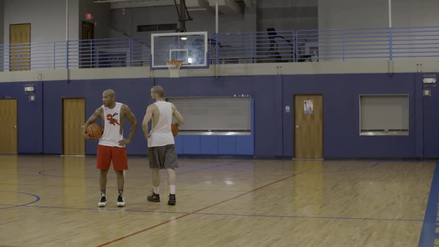 Two men hold and dribble basketballs on an interior basketball court
