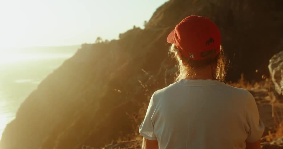 A person wearing a red cap stares at the ocean from the side of a cliff