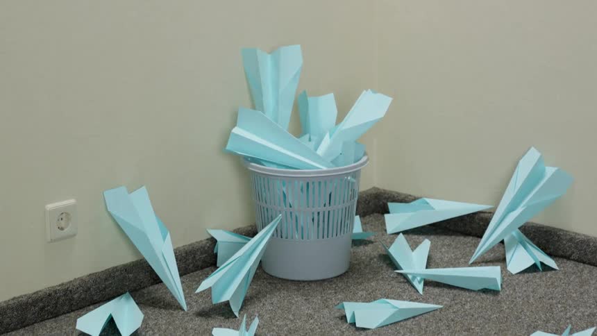 Small waste basket in corner of room with many paper planes in and around it